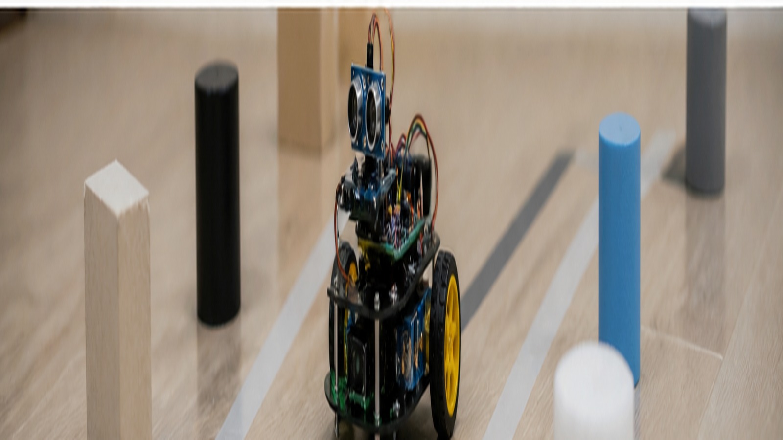 Small wheeled robot prototype with distance sensors and wires navigating around simple test objects.
